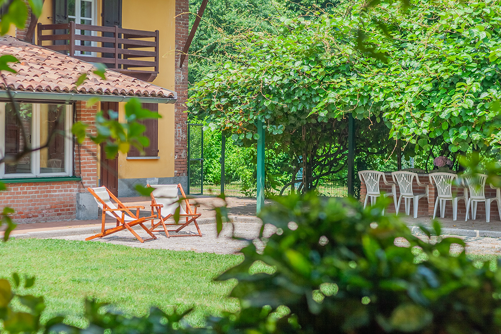 Soggiorno luminoso arredato in stile moderno e minimale, con divano grigio, tavolino in legno, poltrona intrecciata e piante verdi; grande finestra con accesso al balcone e vista panoramica sulle colline.
