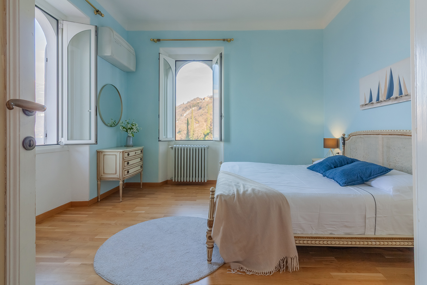 Soggiorno luminoso arredato in stile moderno e minimale, con divano grigio, tavolino in legno, poltrona intrecciata e piante verdi; grande finestra con accesso al balcone e vista panoramica sulle colline.